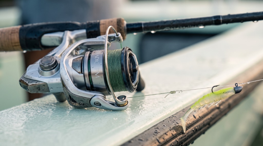 Macro detail of Shimano Stradic FM spinning reel mounted on inshore rod with braided line, FG leader knot, and rigged paddletail swimbait on jig head resting on skiff boat gunwale